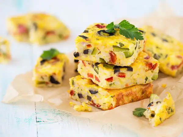 Carrés de frittata colorés, une recette avec des oeufs, garnie de légumes et d'olives, sur du papier sulfurisé.