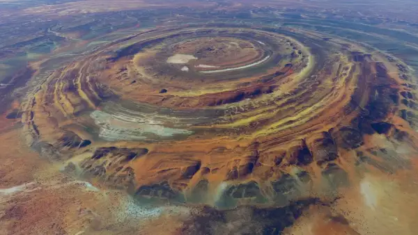 Ökenlandskap med ringformade formationer, som ett gigantiskt öga i Sahara. Färger av orange, gult och blått skapar ett surrealistiskt mönster.