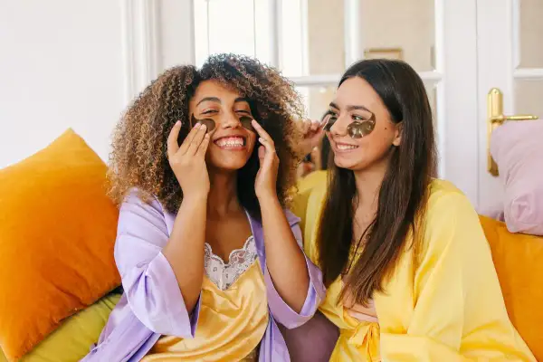 Due amiche sorridono mentre applicano patch per gli occhi, un essenziale skincare step, indossando vestaglie colorate.