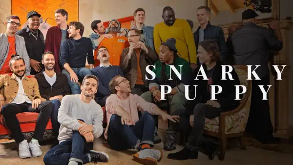 Grupa ludzi, w tym mężczyzna w pomarańczowym kombinezonie, pozuje do zdjęcia z napisem "SNARKY PUPPY".