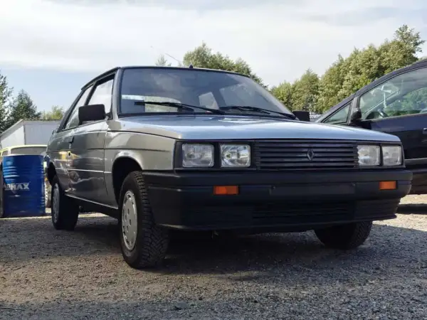 Srebrny Renault 11 z lat 80. stoi na żwirowym podjeździe, obok niebieskiego beczki i innego samochodu.