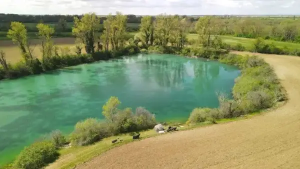 Idyllischer Paylake in Frankreich mit türkisfarbenem Wasser, umgeben von grüner Vegetation und Feldern. Angler haben ihr Lager aufgeschlagen.