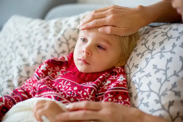 Małe dziecko w czerwonym sweterku z wzorem płatków śniegu leży chore, z gorączką. Ręka opiekuna sprawdza temperaturę. To typowy obrazek, gdy dopada nas wirus.