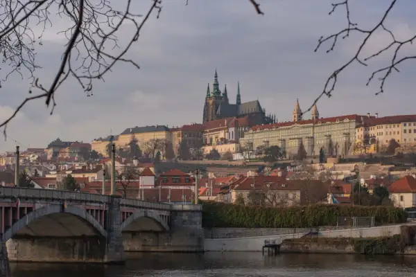 Praga nietypowe atrakcje: widok na Hradczany i Most Karola przez Wełtawę, z gałęziami drzew w pierwszym planie.