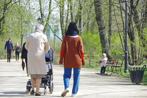 Kobieta z wózkiem spaceruje po parku, w tle inne osoby i dziecko na ławce.