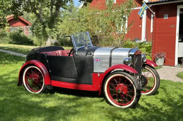 En klassisk Austin bilmodell i svart och rött med brittisk flagga, parkerad på en gräsmatta framför ett rött hus med svensk flagga.