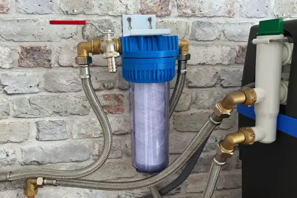 Filtre à eau avec cartouche visible, raccordé à un adoucisseur qui coule en permanence. Tuyaux flexibles et robinet rouge.
