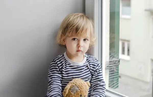 Smutny dwulatek nie mówi, tuli misia przy oknie.