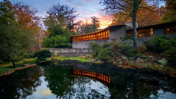 The last Wright house, a modern masterpiece, reflects warmly in a tranquil pond amidst autumn foliage.