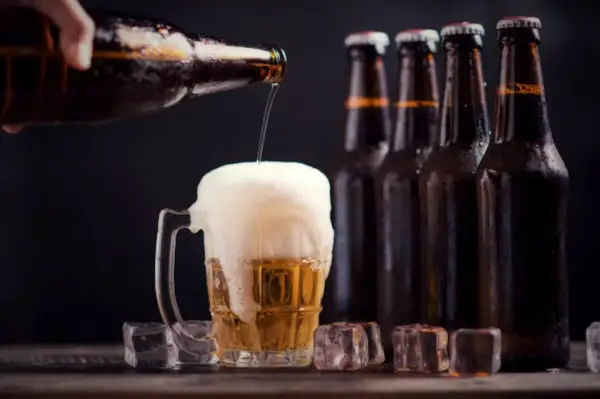 Una mano versa birra in un bicchiere traboccante di schiuma. Sullo sfondo, bottiglie di ibu birra e cubetti di ghiaccio.