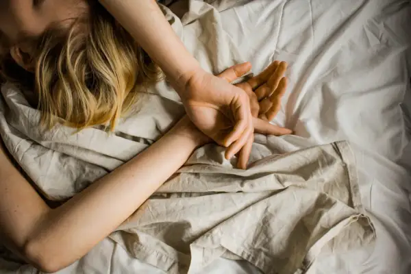 Blonde Haare auf einem Kissen, Arme schlafen nachts ein, Hände ruhen auf der Brust, umgeben von zerknitterter Bettwäsche.