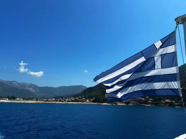 Flaga Grecji powiewa nad błękitnym morzem, w tle widać górzyste wybrzeże z zabudowaniami. Idealny dzień na plażowanie w Grecji.