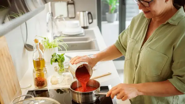 Mujer vertiendo tomate triturado en una olla para espesar una salsa. Ingredientes como aceite, ajo y especias a la vista.