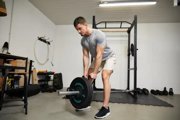 Homme musclé en buste penché, soulevant une barre chargée de poids dans une salle de sport à domicile.
