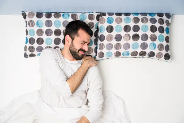 Uomo a letto con dolore alla spalla, dormire con le braccia alzate non è comodo.