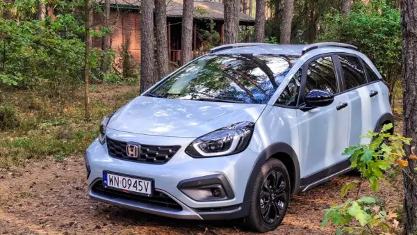 Jasny Honda Jazz Crosstar zaparkowany w lesie, obok drewnianego domku.