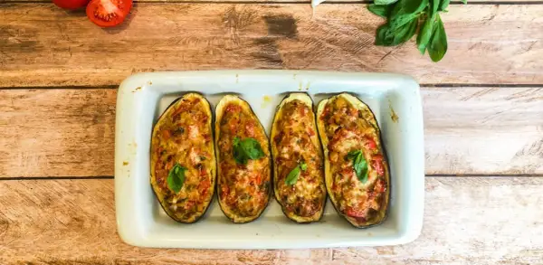 Quatre moitiés d'aubergines farcies, gratinées et garnies de basilic, dans un plat blanc sur fond de bois.