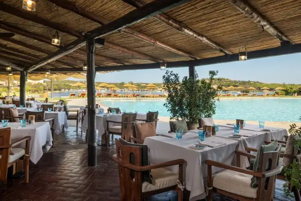 Tavoli apparecchiati sotto un tetto di paglia, con vista sulla piscina e sulla spiaggia del **hotel Cala di Volpe**.