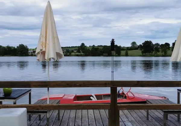 Gemütliches Restaurant am See mit rotem Tretboot und Sonnenschirmen. Die Uferlandschaft ist grün und hügelig.