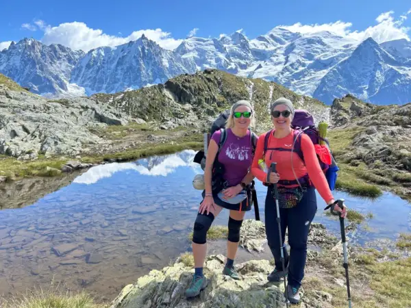 Dwie kobiety cieszą się trekkingiem wokół Mont Blanc. Mają plecaki i kije trekkingowe, a w tle widać ośnieżone szczyty.