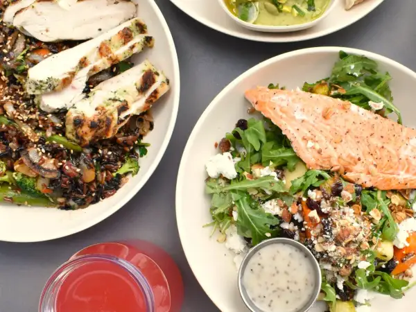Två skålar med proteinrik mat: grillad kyckling med vildris och grönsaker, samt lax med sallad, nötter och ost. En röd dryck syns också.