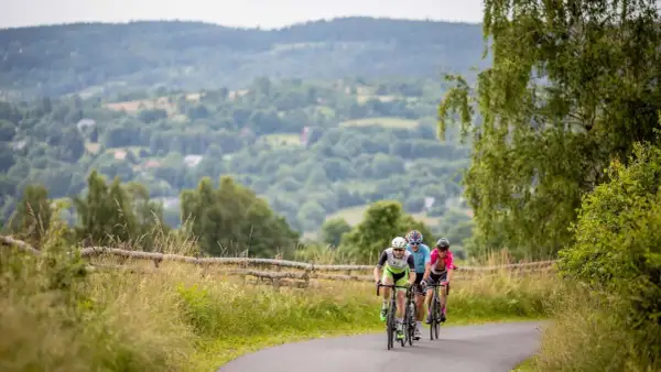 Trzech rowerzystów jedzie po asfaltowej drodze wśród zielonych wzgórz.
