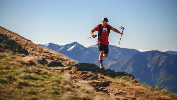 Ein Mann im roten Shirt und schwarzer Hose läuft mit Stöcken einen Bergpfad entlang. Sein intensives anaerobes Training fordert ihn in der atemberaubenden Berglandschaft.