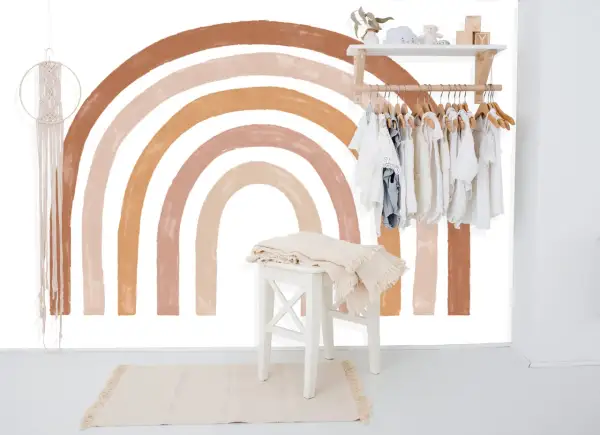 Un arc-en-ciel aux tons terreux est peint sur un mur blanc, orné d'une penderie d'enfants et d'une décoration murale en macramé.