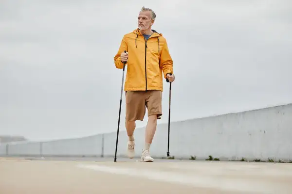 Mężczyzna w żółtej kurtce i brązowych spodenkach wykonuje rozgrzewkę przed nordic walking, trzymając kije.