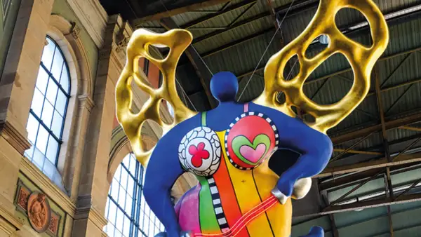 Silhouette d'une Nana colorée de Niki de Saint Phalle, avec des ailes dorées, suspendue dans un grand hall.