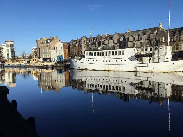 Barcos y edificios históricos reflejados en el agua en Leith, Edimburgo. El "Ristorante De Niro" en un barco blanco destaca.