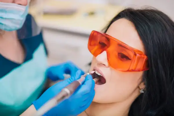 Pacjentka w okularach ochronnych przechodzi zabieg znieczulenia komputerowego zęba. Dentysta w rękawiczkach używa nowoczesnego urządzenia.