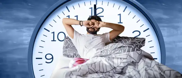 Man in bed rubs his eyes, looking tired. A large clock face behind him shows it's just past 6 AM, suggesting only six hours of sleep.