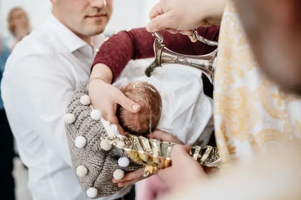 Chrzest dziecka podczas chrzcin. Woda z ozdobnego dzbanka spływa na główkę niemowlęcia.