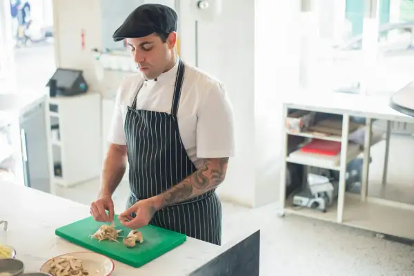 Chef con gorra y delantal a rayas, cortando champiñones en una tabla verde. Ideal para aprender a cocinar.