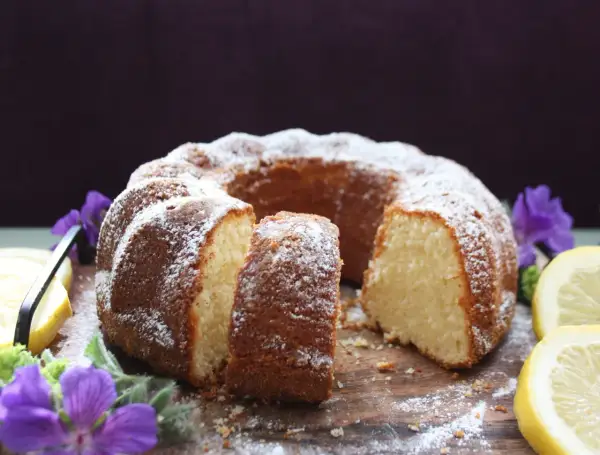 En saftig citron sockerkaka, pudrad med florsocker, serveras med citronskivor och lila blommor på en träbricka.