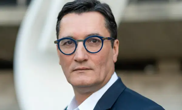Olivier Dartigolles, un homme aux lunettes bleues, pose pour un portrait. Il est vêtu d'un costume sombre, évoquant une image de famille et de professionnalisme.