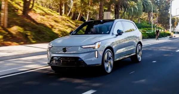 Szary Volvo EX90 jedzie po słonecznej drodze, otoczony zielenią i palmami.