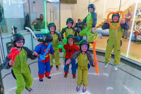 Niños con cascos y trajes verdes disfrutan de experiencias únicas, algunos con globos de colores, en un túnel de viento.