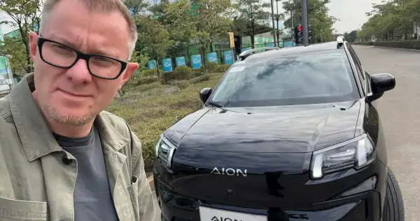 Mężczyzna w okularach pozuje do selfie obok czarnego SUV-a marki AION, jednej z nowych marek samochodów.