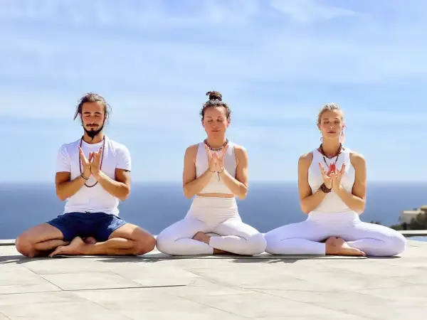 Tres personas meditan al aire libre, con las manos juntas en un mudra. El mar y el cielo azul forman el telón de fondo.