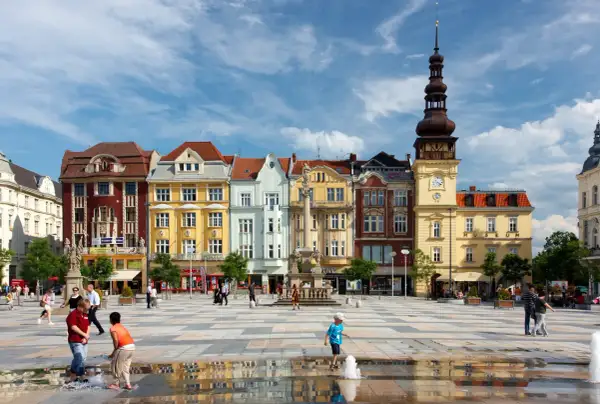 Kolorowy plac w Ostrawie z ludźmi i zabytkowymi budynkami w tle.
