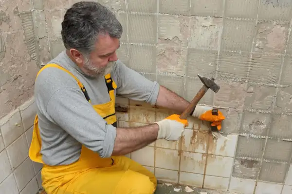 Homme en salopette jaune en train d'enlever du carrelage mural avec un marteau et un burin.