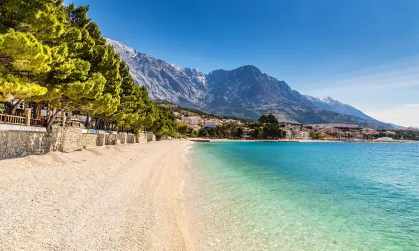 En vacker strand med turkost vatten och en bergskedja i bakgrunden. En promenadstråk kantas av träd och stenmurar, perfekt för en avkopplande stund vid makarska strandpromenad.