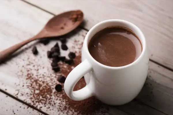 Tazza di cioccolata calda con cacao amaro, gocce di cioccolato e un cucchiaio di legno su un tavolo di legno.