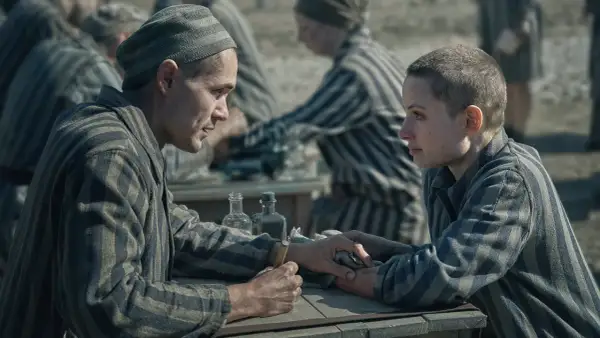 Två fångar i randiga uniformer möts vid ett bord. Mannen håller en nål, redo att tatuera kvinnan. Scen från filmen "Tatueraren i Auschwitz".