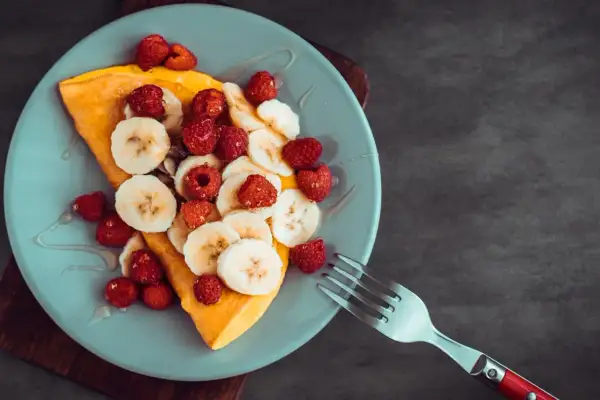 Omlet z bananem i malinami, polany syropem, na jasnoniebieskim talerzu. Obok leży widelec.