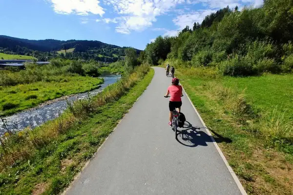 Rowerzyści przemierzają malowniczą trasę velo Krynica wzdłuż rzeki, otoczeni zielonymi wzgórzami i lasami.