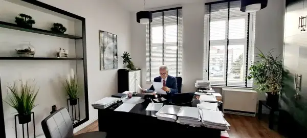 Mężczyzna w garniturze siedzi przy biurku w gabinecie, przeglądając dokumenty.