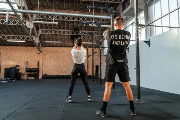 Para osiągnięcia dobrych wyników w crossfit, jak często trenować? Para ćwiczy w siłowni, kobieta z kettlebell, mężczyzna w koszulce "It's Going Down".
